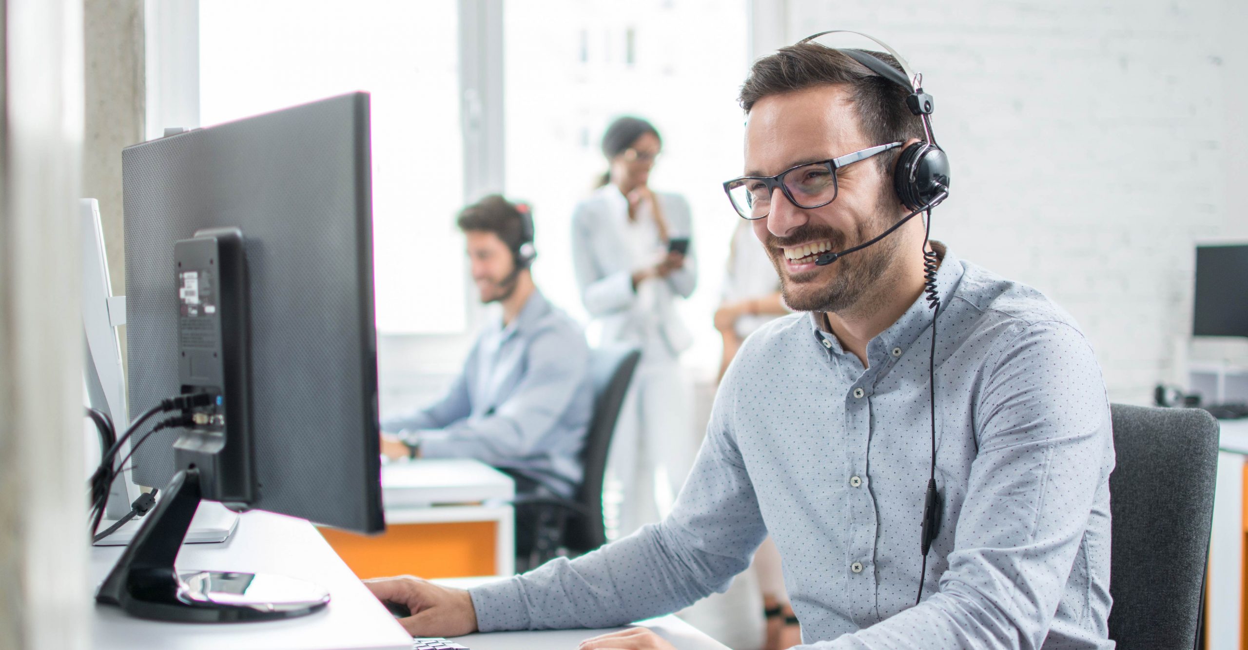 A man wearing a headset and glasses sits at a desk with a computer monitor, smiling. Three other people are in the background, also working at desks.