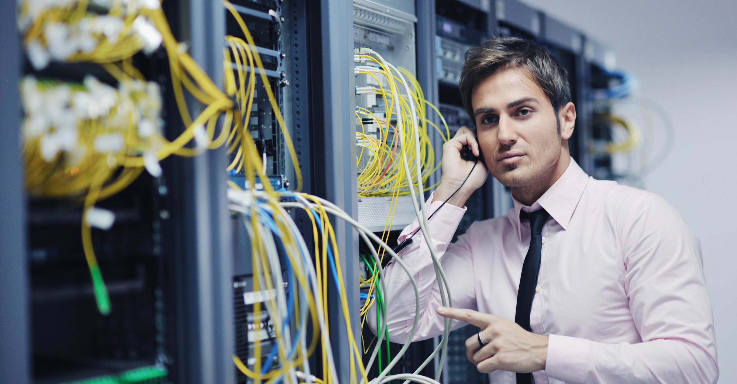 Man in a white shirt and tie stands in a server room, holding a cable and pointing to network equipment with various colored cables connected to it.