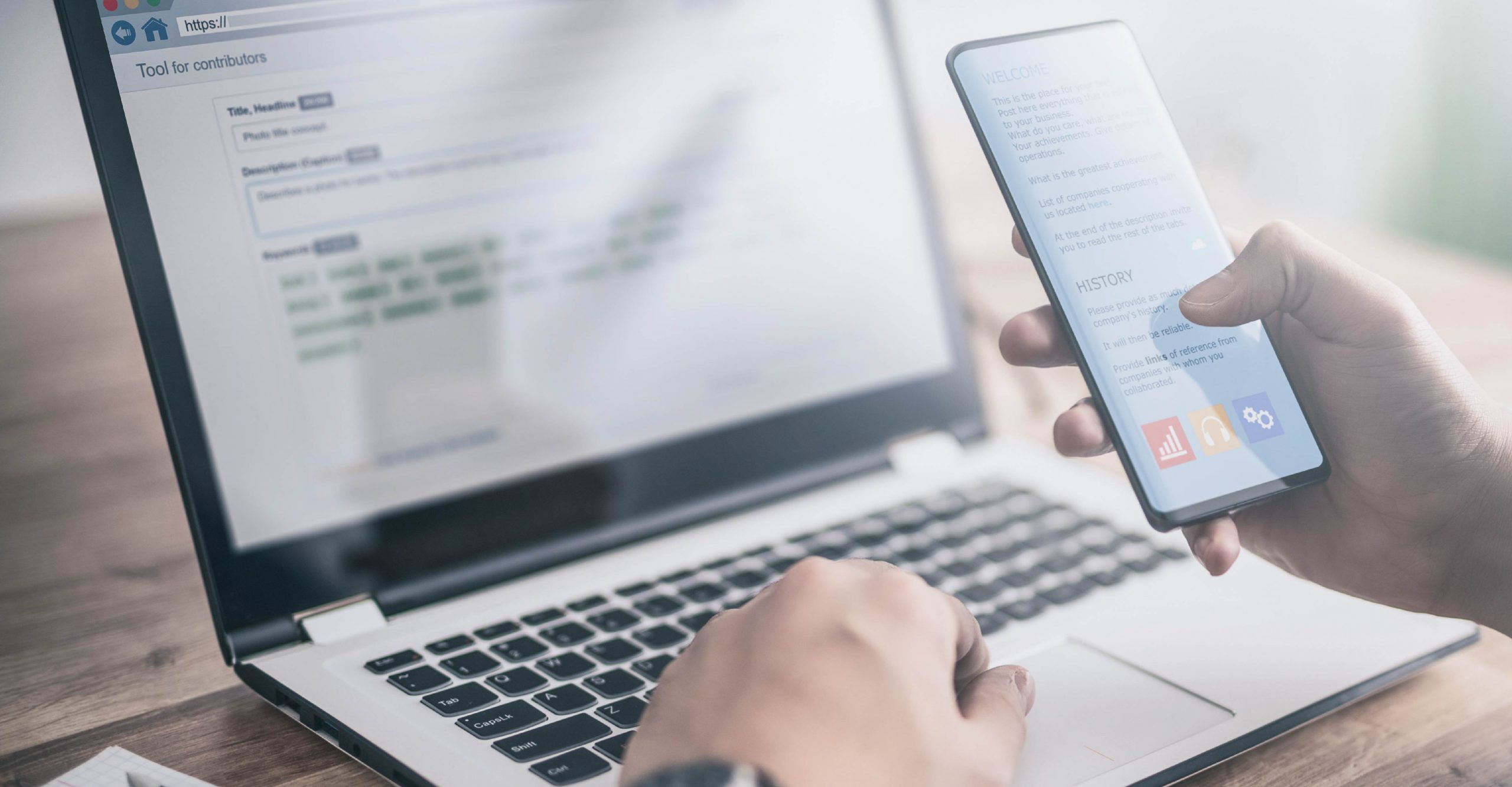 Person using a smartphone and laptop simultaneously; the smartphone displays an app interface, and the laptop screen shows a form or text editor.