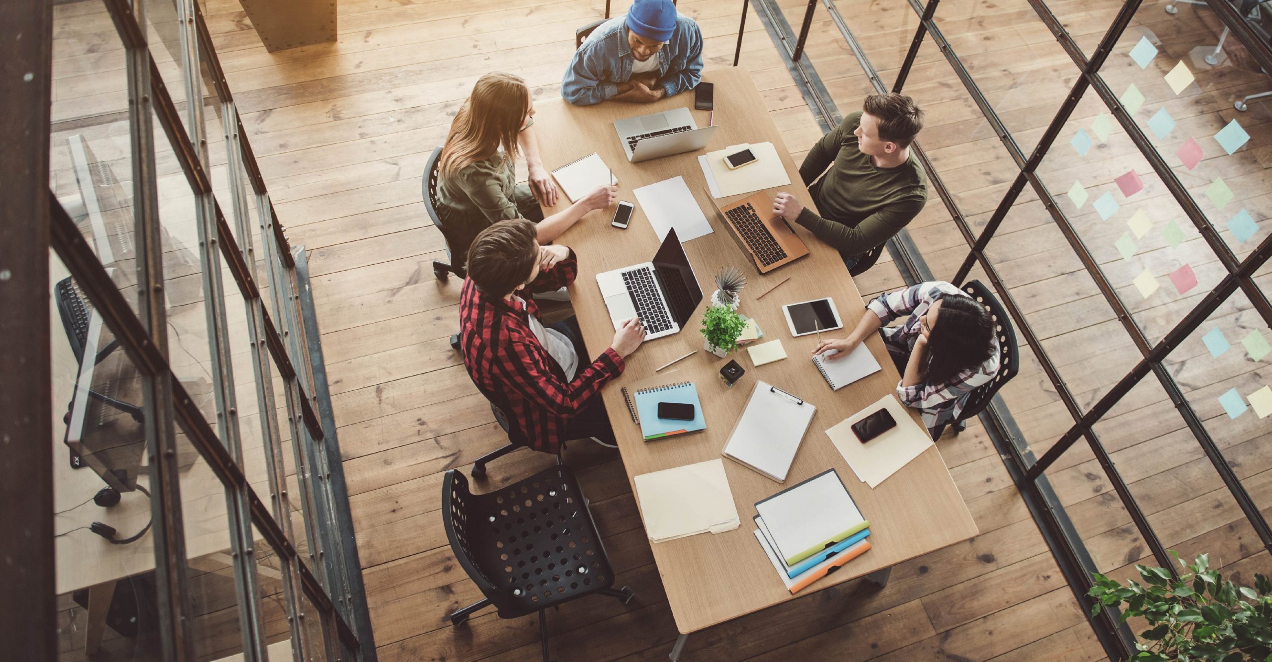 Five people sit around a table in an office, working on laptops and using notebooks. The room has a wooden floor and large glass windows. Various office supplies and a small plant are on the table.