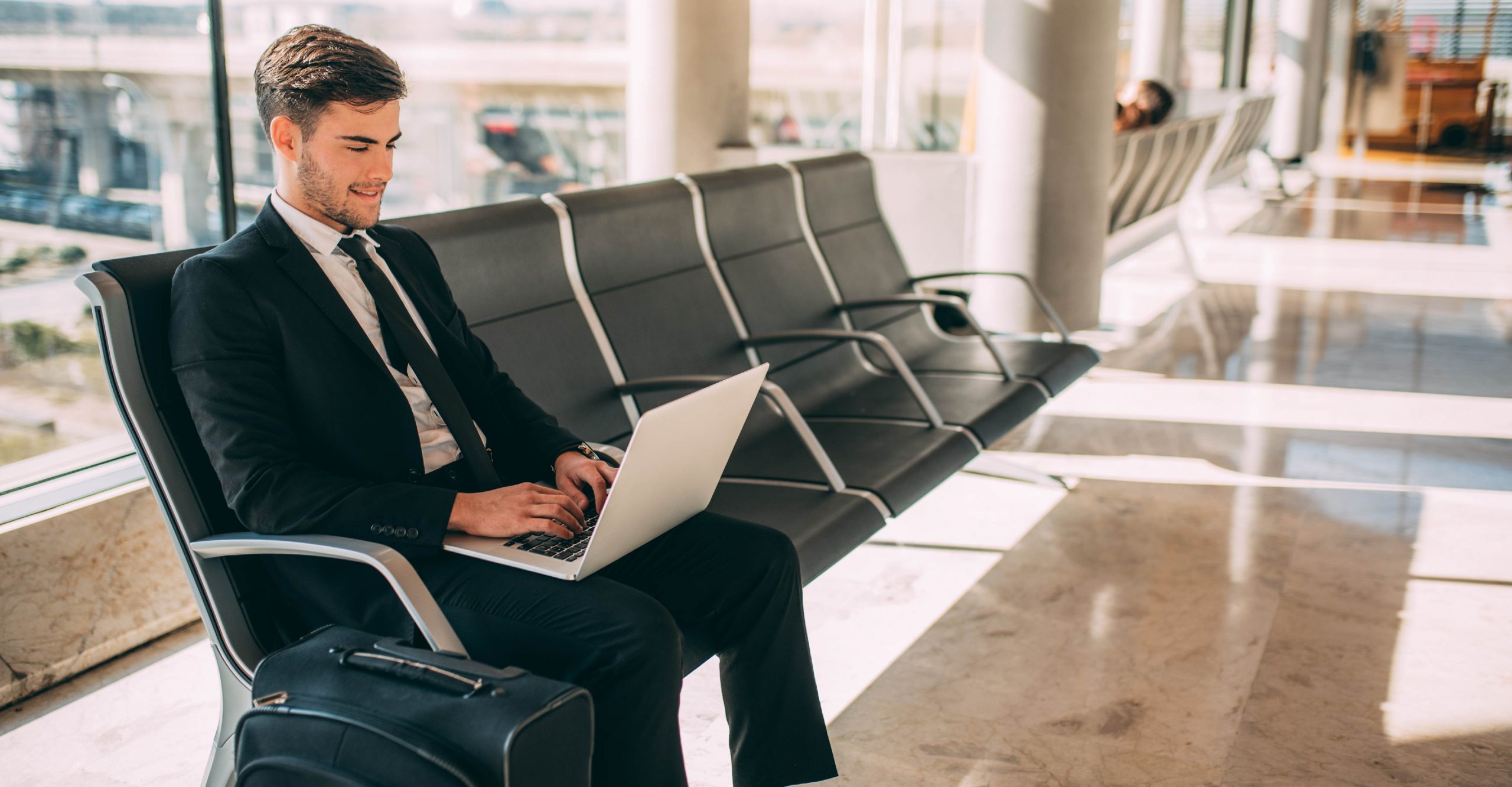 A man in a suit sits in an airport lounge, typing on a laptop with his suitcase next to him.