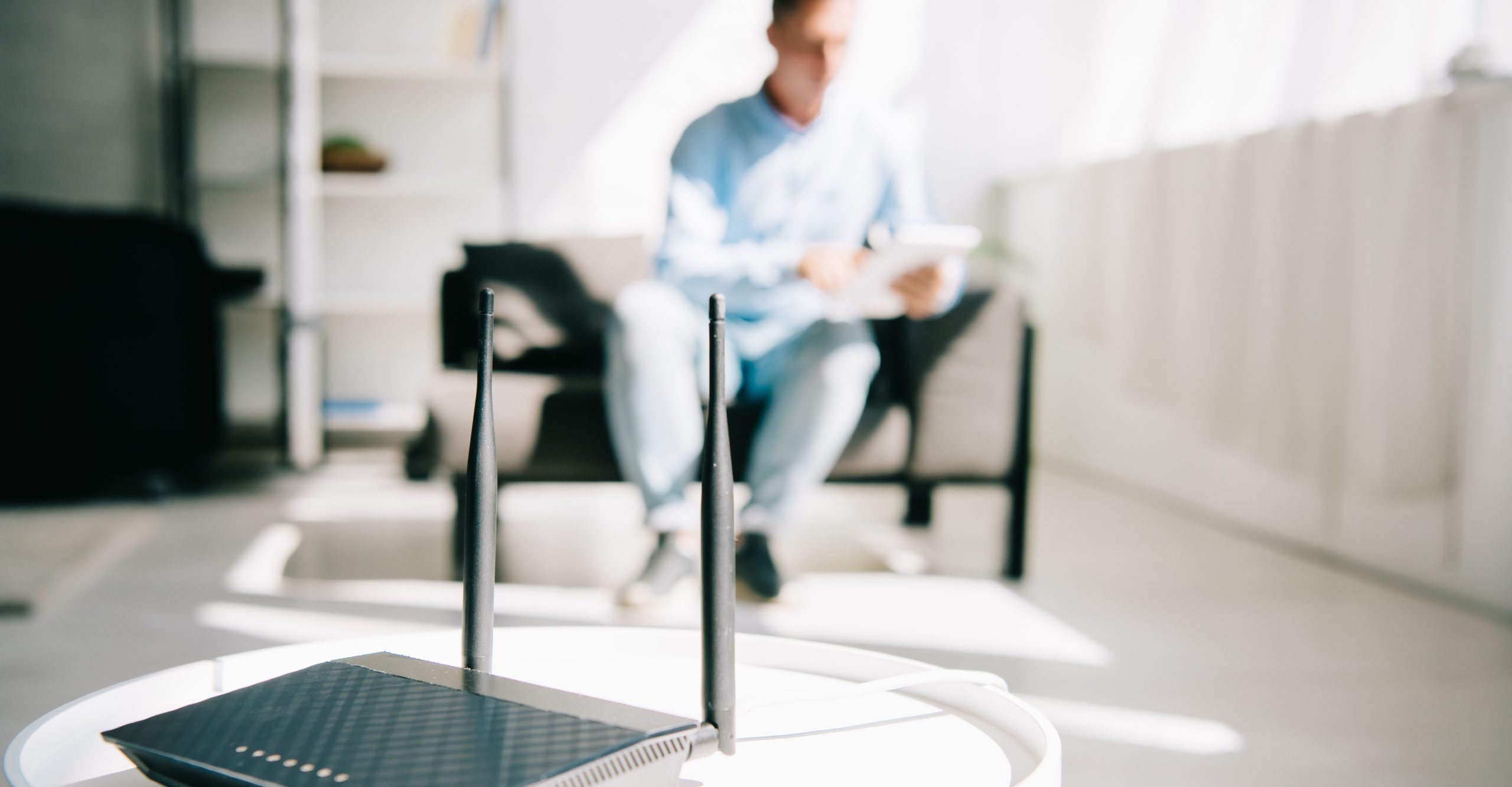 A black Wi-Fi router with two antennas sits on a white table, its LED lights blinking. In the background, a person relaxes on the couch in a bright room, using a tablet secured with a strong password.