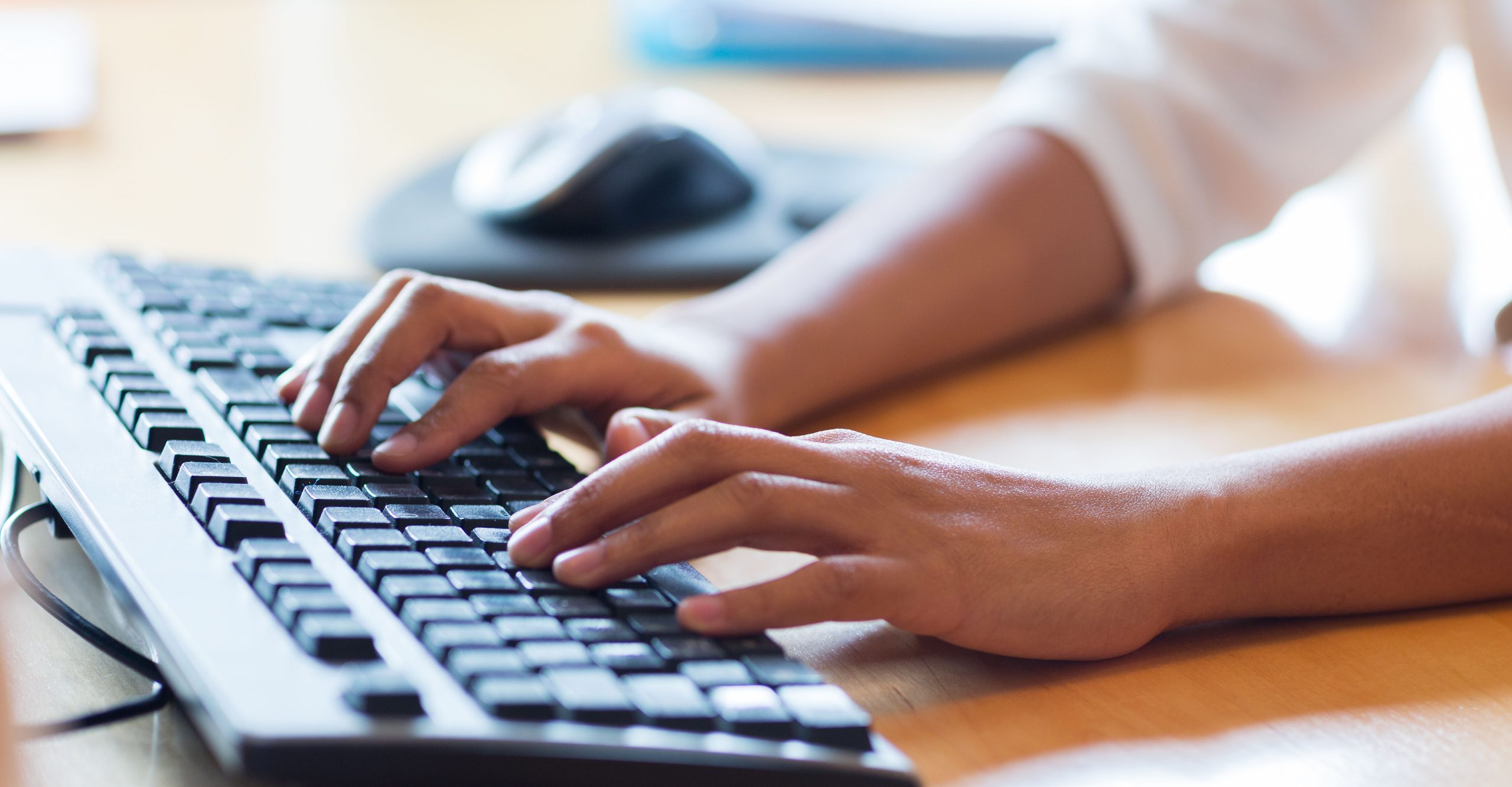 Hands typing a password on a computer keyboard with a mouse nearby on a wooden desk.