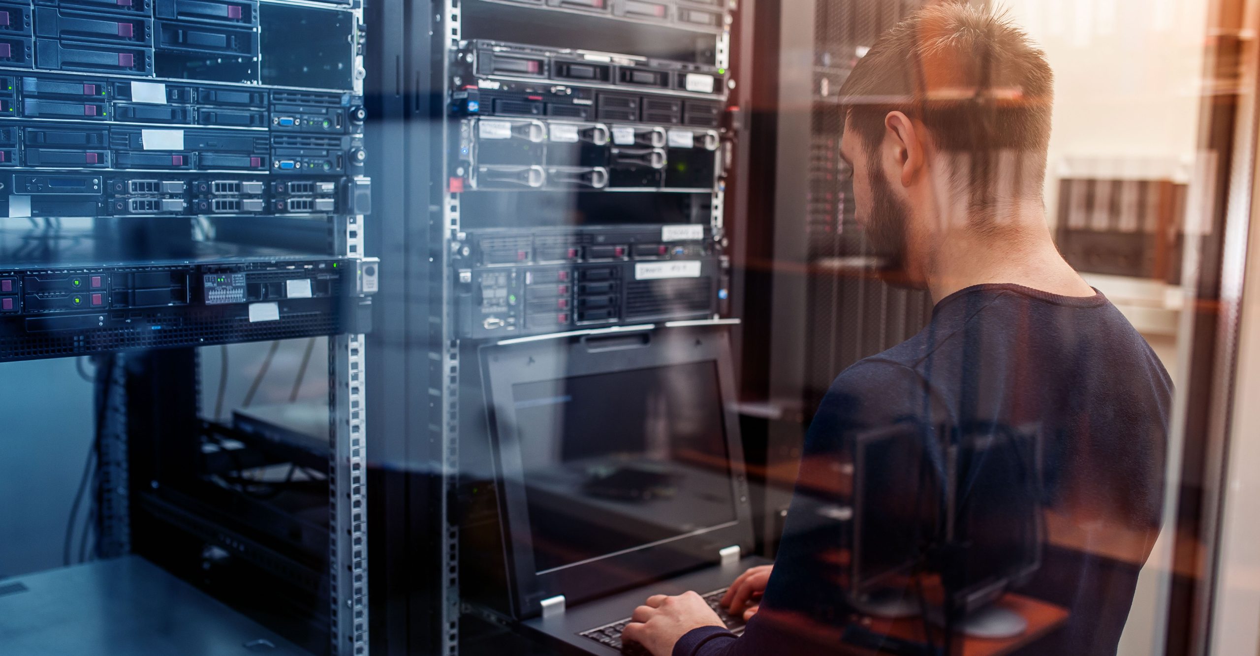 A person focused on their laptop in a server room, surrounded by racks of computer hardware, diligently handles endpoint management.