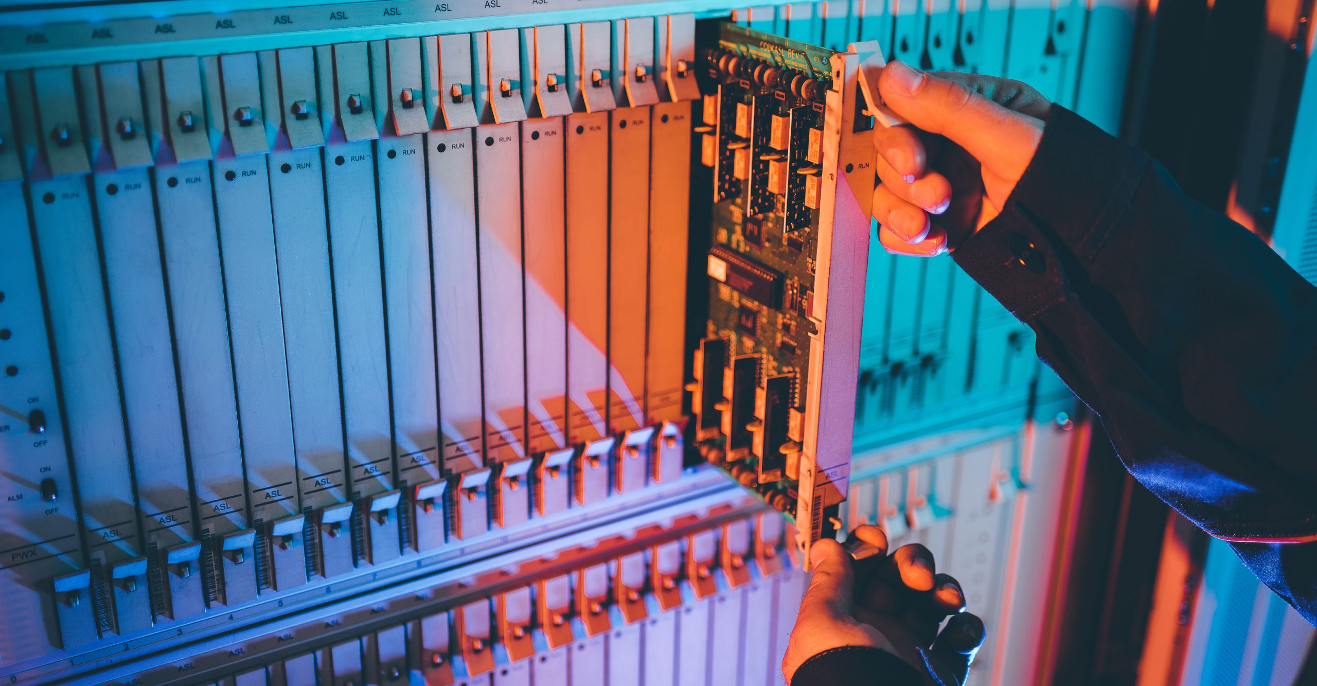 Hands carefully inserting a circuit board into a rack of electronic components, illuminated with blue and orange lighting, exemplifying the precision and expertise required in server management.