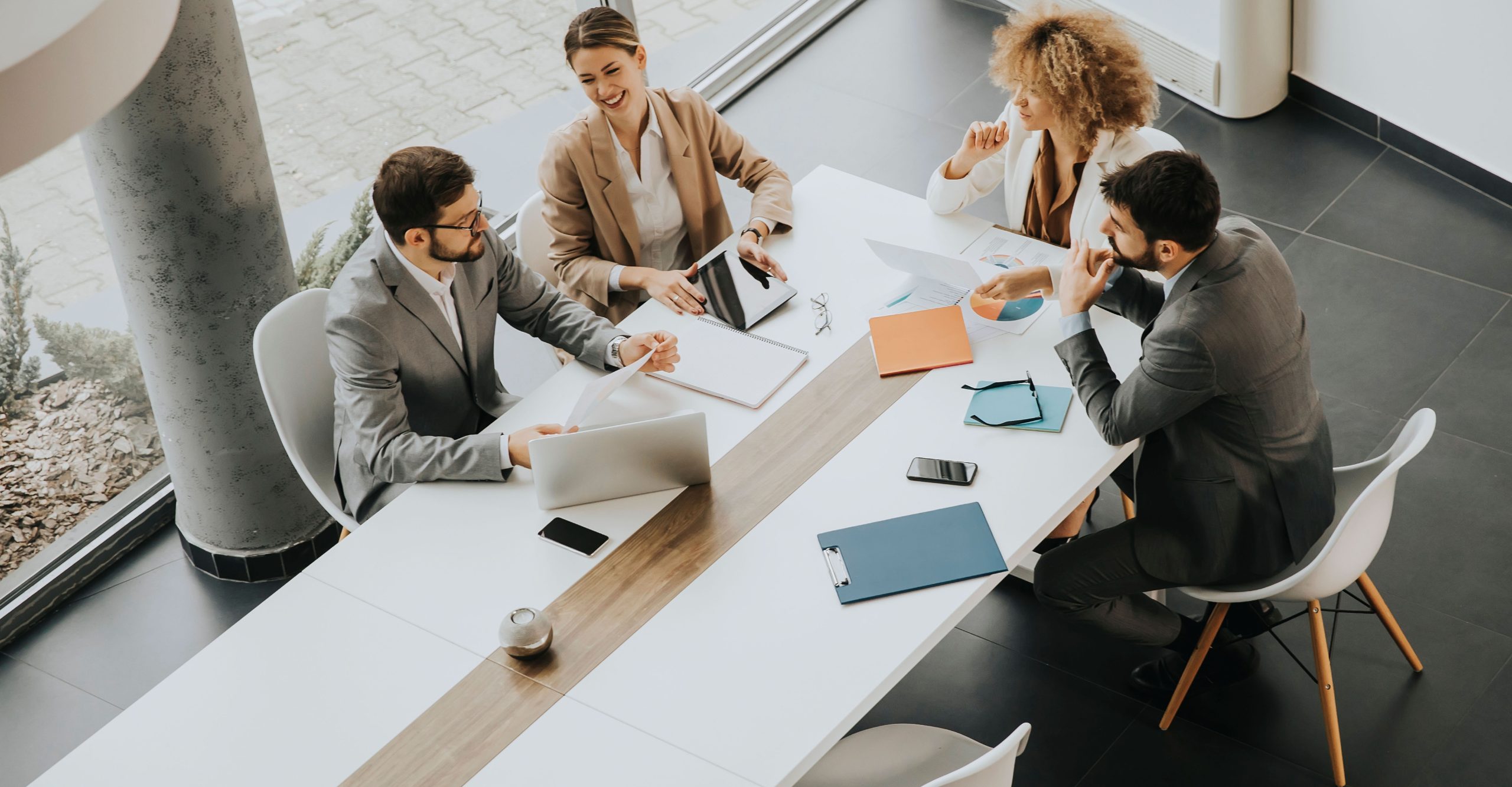 Four professionals are seated around a conference table in an office setting, engaged in a discussion about EDR vs. MDR strategies. Laptops, tablets, documents, and coffee cups are on the table. Large windows provide natural light.