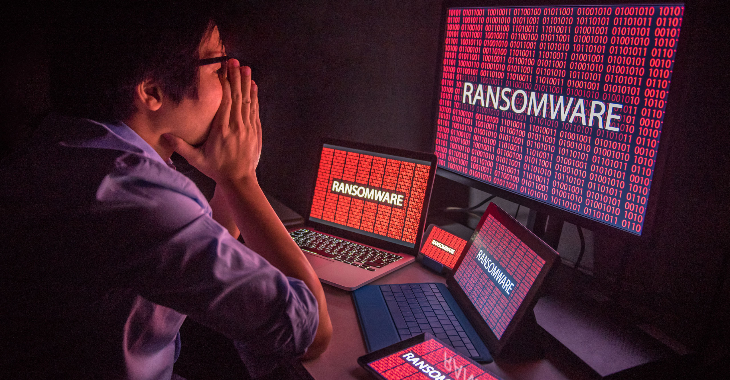 A person sits at a desk with multiple screens displaying the word "RANSOMWARE" and lines of binary code in red, suggesting a cybersecurity threat. The scene highlights the urgent need for systems hardening to defend against such attacks.