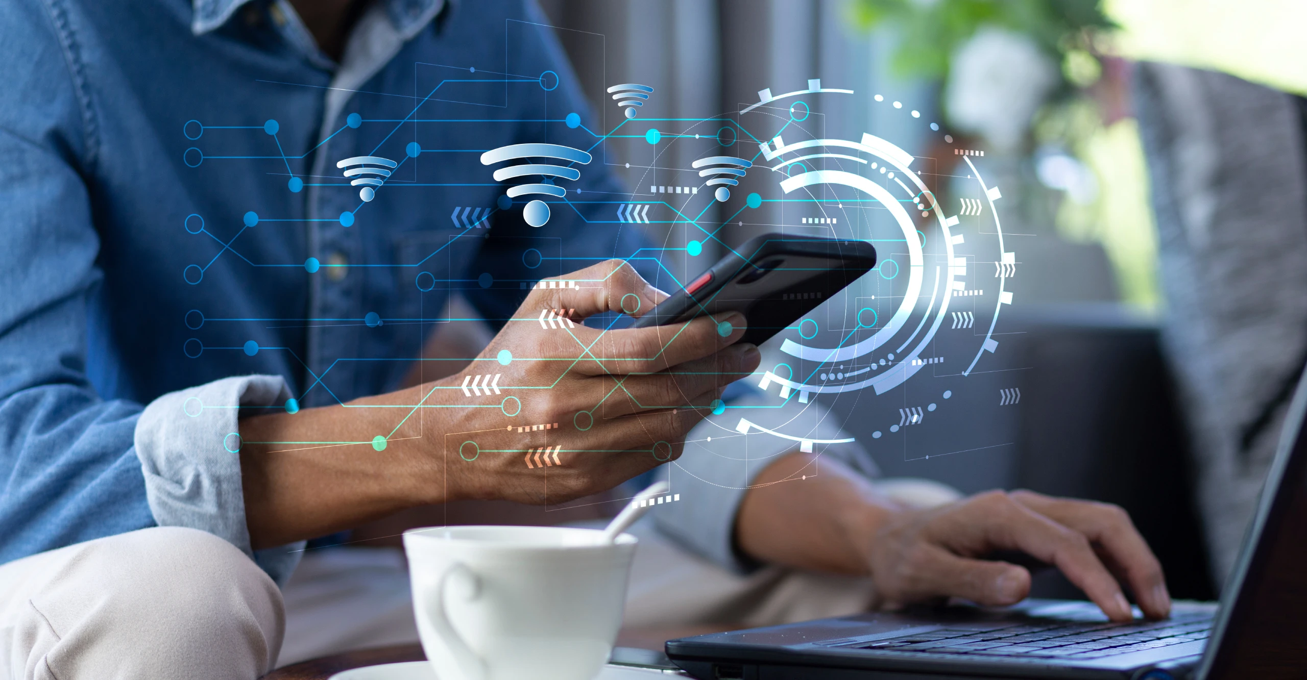 A person holds a smartphone displaying digital icons while typing on a laptop, engaging in systems hardening. A cup of coffee is in the foreground.