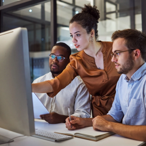 Three people collaborate in an office focusing on a large computer screen. One person, seemingly from an IT support company, gestures towards the screen while explaining something to the others, who take notes and listen attentively.