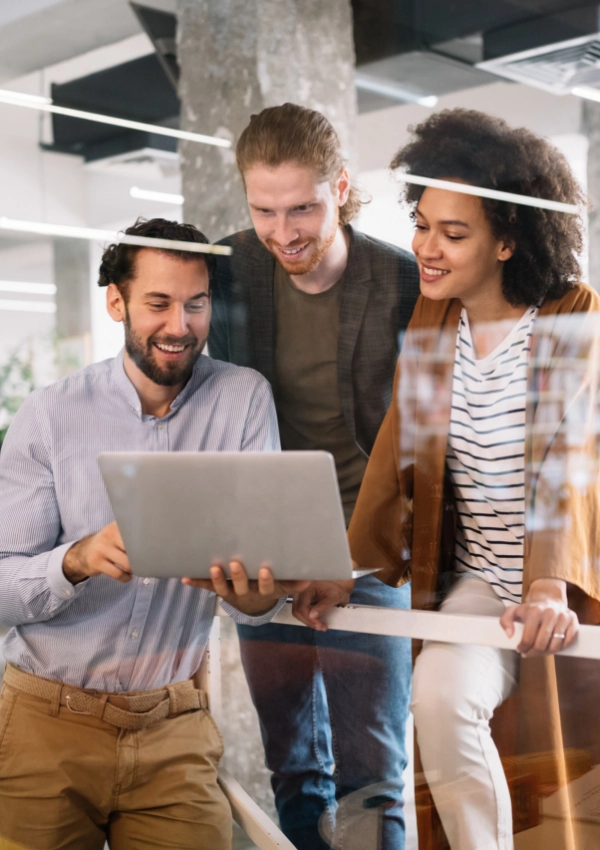 Three colleagues stand together in an office, looking at a laptop and smiling, as they explore new solutions with the help of their IT support company.