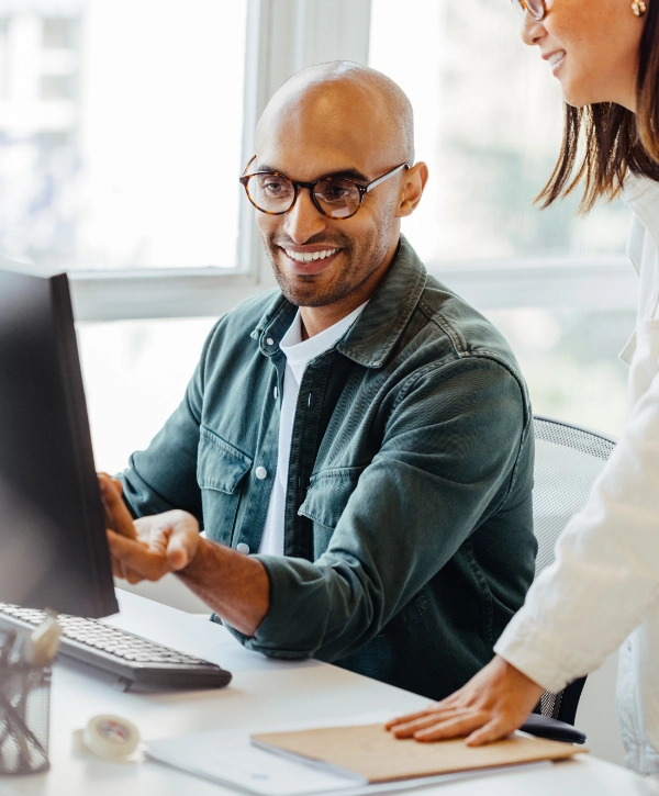 A smiling man wearing glasses works at a computer while a woman, also smiling, stands beside him. Both appear to be discussing managed IT services on the screen in a bright office environment.