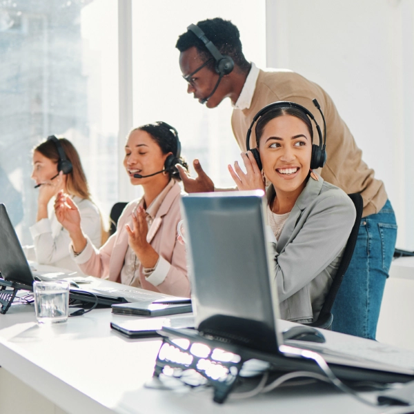 Four call center agents wearing headsets work at their computers in a brightly lit office, engaging in conversation and smiling as they provide computer IT support to clients.