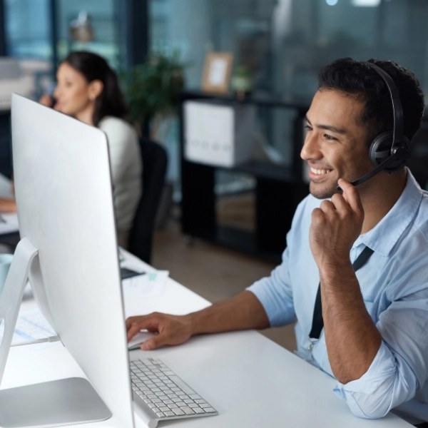 A man is sitting at a desk wearing a headset and smiling while providing IT support on a desktop computer. Another person is visible in the background.