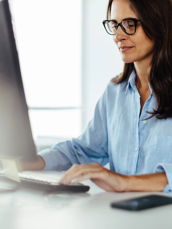 A woman wearing glasses and a blue shirt sits at a desk, focused on her computer screen, providing diligent network support.