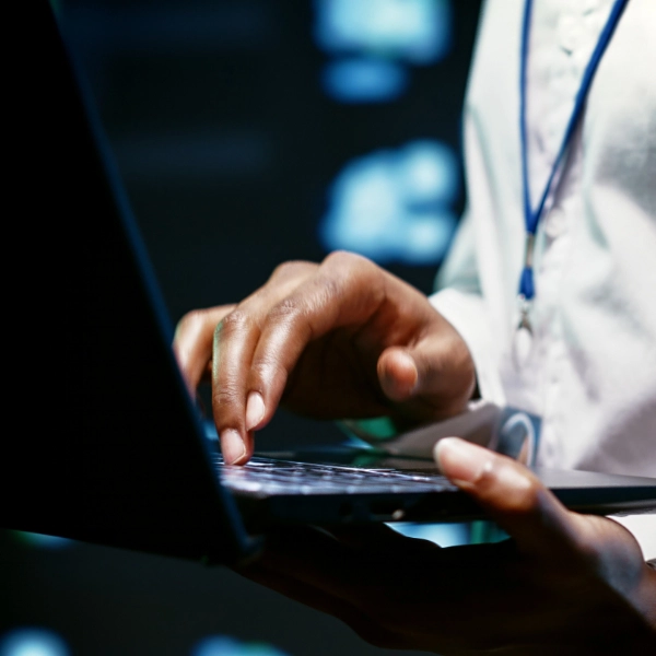 A person wearing a white shirt and a lanyard is using a laptop with both hands in a dimly lit environment, representing the meticulous dedication often seen in an IT support company.