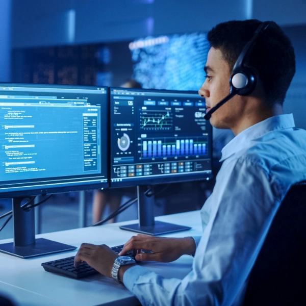 A person wearing a headset is sitting at a desk with two monitors displaying technical data and graphs. The individual, representing an IT support company, is typing on a keyboard in a modern office environment.