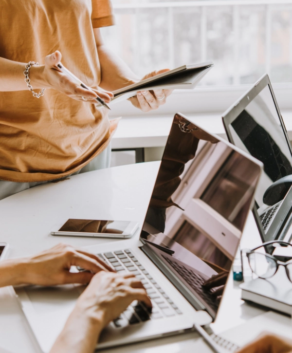 Two people collaborating in an office with laptops and tablets on a white desk. One person is standing and gesturing with a notebook, while the other is typing on a laptop, embodying the essence of managed IT services.