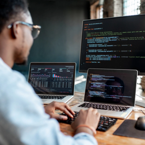 A person wearing glasses works on a computer with three screens displaying code and data analytics in a modern workspace, providing essential IT services.