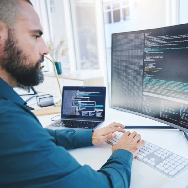 A person with a beard works at a desk with a large monitor and a laptop, both displaying code. They are typing on a keyboard with focused attention, representing the dedication found in an IT support company.