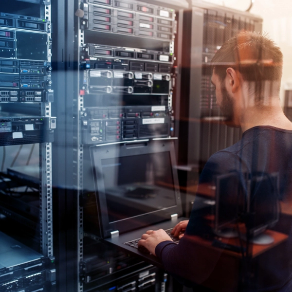 A person works on a laptop in a server room, providing IT support amidst racks filled with servers and networking equipment.