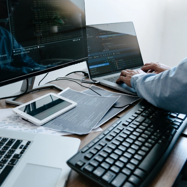 A person working at a desk with multiple screens displaying code, a tablet, and a keyboard, surrounded by papers, providing IT support services.