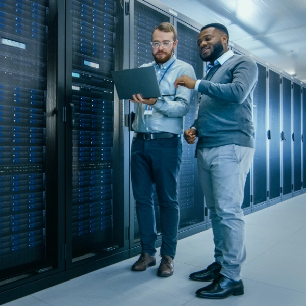 Two men standing in a server room. One, from an IT support company, is holding a laptop while the other points at the screen. Rows of server racks are visible in the background.