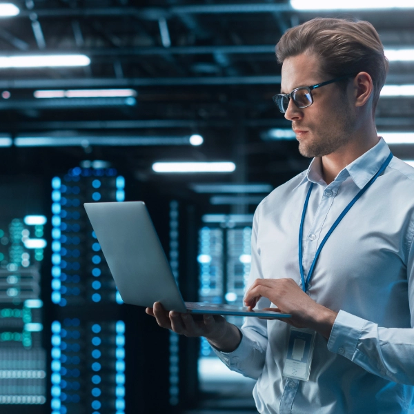 A person wearing glasses and a white shirt stands in a server room, working on a laptop, providing network support. The background shows rows of illuminated server racks.