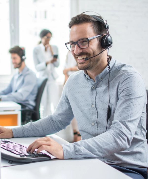 A man wearing glasses and a headset smiles while typing on a keyboard in an IT support company office. Other workers can be seen in the background, showcasing the collaborative environment of managed IT services.