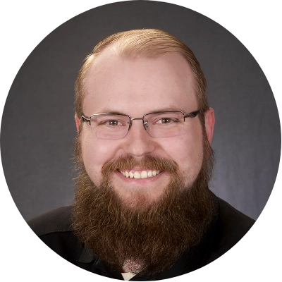 A man with glasses and a thick beard smiling against a dark background.