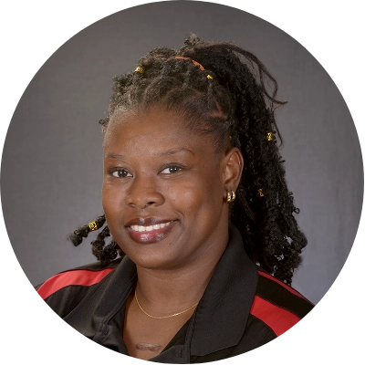 A person with braided hair styled in a ponytail, wearing a black and red collared shirt, smiles against a plain background.