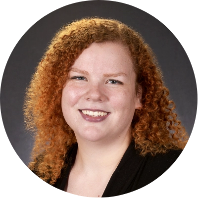 A person with curly red hair and a black top smiles at the camera against a dark background.